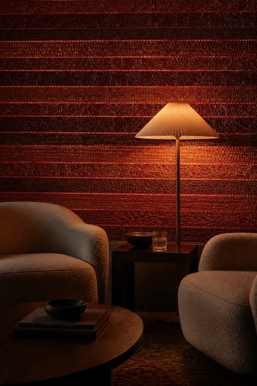 Members club lounge at evening with Garnet Reed burgundy-red grasscloth wallpaper, low bouclé armchairs, dark oak side table, and warm floor lamp